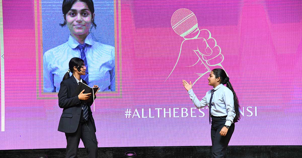 Two individuals on stage presenting with a tablet and microphone against a vibrant background at JIRS.