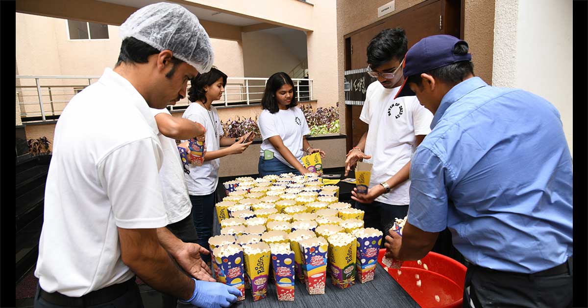 Five individuals prepare popcorn in a lobby area, wearing T-shirts and uniforms, engaged in a group activity.