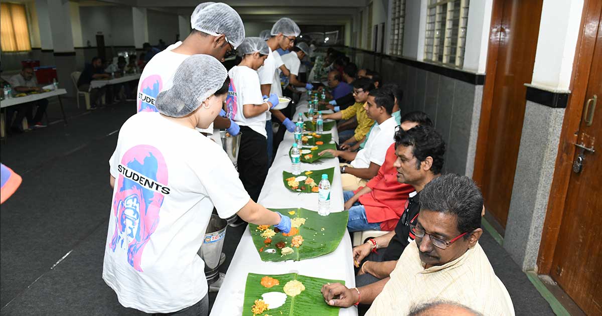 Students serve traditional meals on banana leaves at a communal dining event indoors.